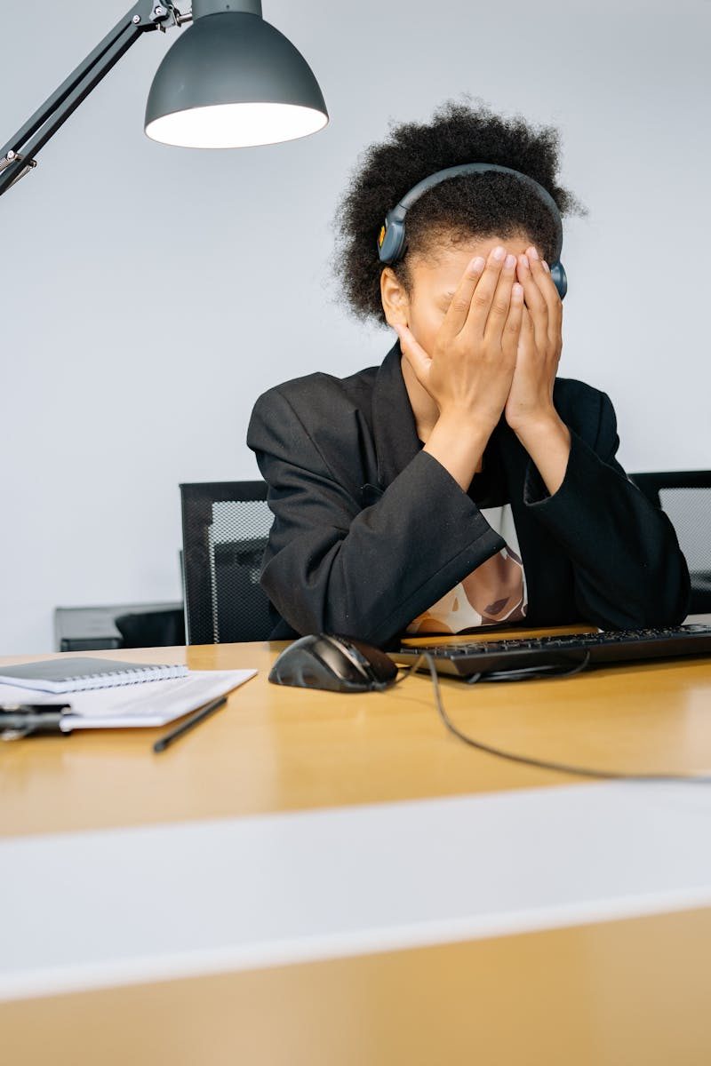 A tired call center employee covering her face with hands in frustration, wearing a headset in an office.